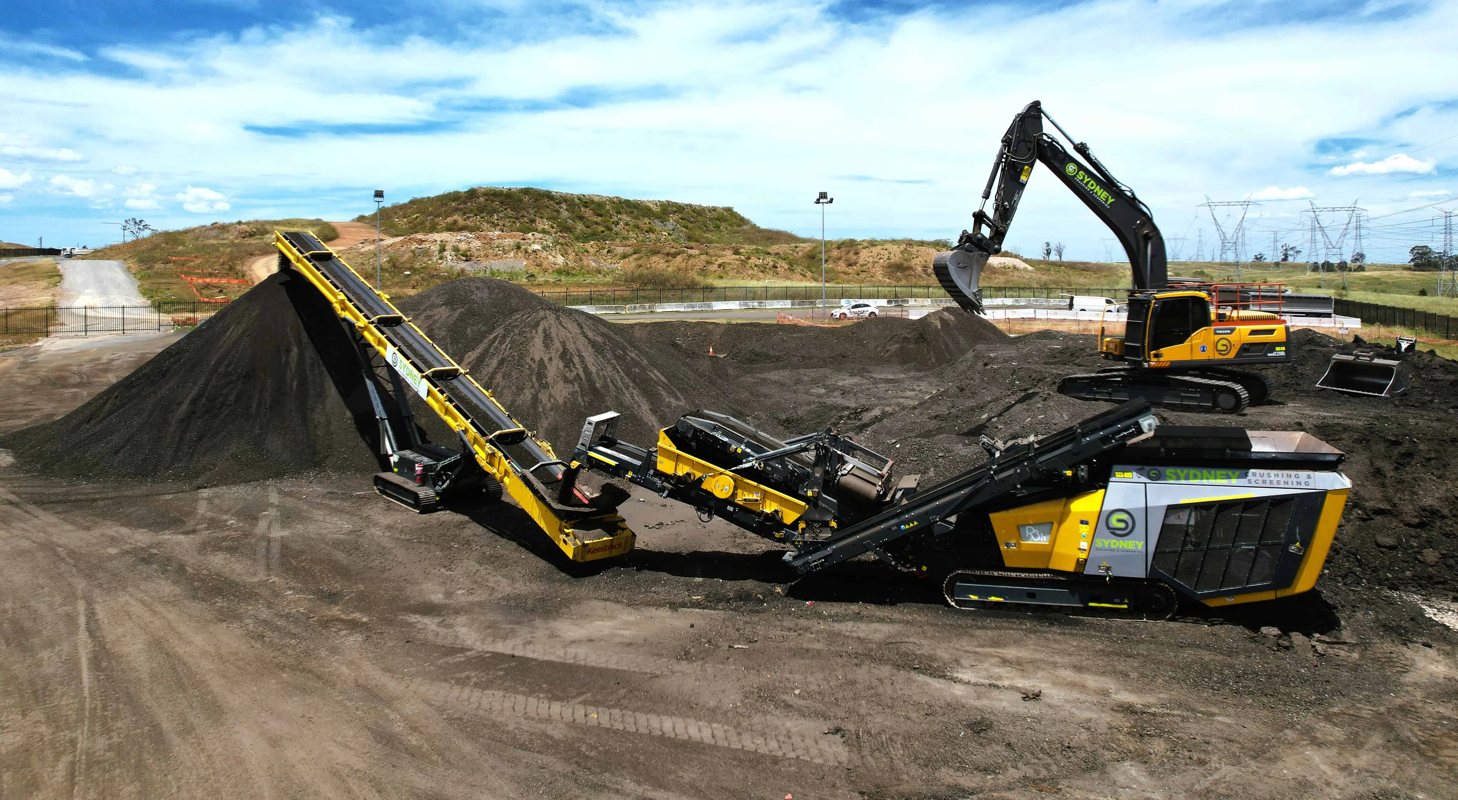 Crusher, conveyor and excavator working a stockpile of waste asphalt on site